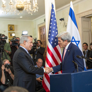 Benjamin Netanyahu and John Kerry shaking hands at a joint press conference. The room is filled with media personnel and officials. American flag prominently displayed.