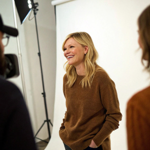 Kirsten Dunst in a candid shot, dressed in a casual brown sweater. Bright white studio backdrop. Her friendly smile is the focal point.