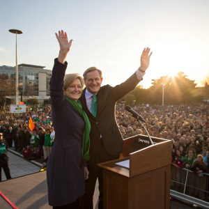 Enda Kenny and Fionnuala Kenny waving from a podium. Large crowd gathered outdoors, sunny day.