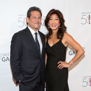 Neal Shapiro and Hyunju 'Juju' Chang at gala event. Shapiro in dark suit, Chang in black dress. White backdrop with event details