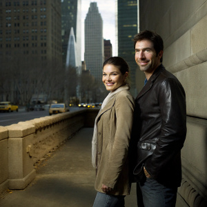 Dylan McDermott and Alexa Davalos, actors from FBI Most Wanted, posing outdoors. Urban landscape with modern skyscrapers and bustling city streets in the background. They are standing closely, smiling.