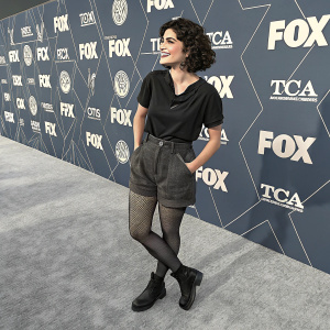 Eve Harlow at a TCA and FOX event. Wearing casual attire: black top, shorts, stockings, and dark shoes. On grey carpet with event logos in the background.