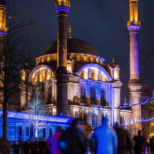 At night, the Ortakoy Mosque in Istanbul, Turkey, is brilliantly lit, casting a warm glow. The imposing structure, with its multiple domes, arches, and ornate details, stands out against the dark sky. The foreground teems with blurred figures, adding a sense of movement and life to the scene.
