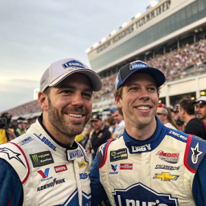 Jimmie Johnson and Brad Keselowski, in racing gear, at NASCAR celebration. Background features large crowd and grandstands.