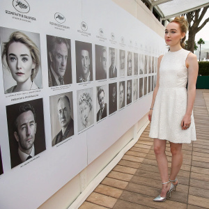 Saoirse Ronan at Cannes Film Festival, standing beside wall of celebrity portraits. She's wearing a simple white dress with delicate lace, paired with silver heels. Her hair is in a loose bun, face serene.
