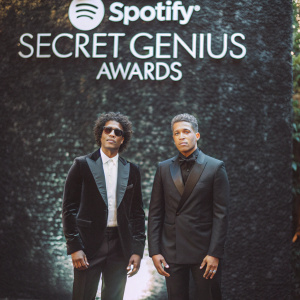 J Kash and Babyface at the Spotify Secret Genius Awards. Both are dressed in black suits and standing in front of a Spotify logo backdrop.