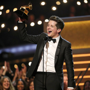 Charlie Puth, dressed in a tuxedo, excitedly accepting an award at the Grammy Awards. Bright stage lights and cheering crowd in background.
