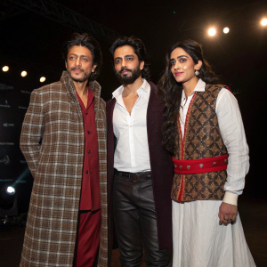 Shah Rukh Khan, Saahil Prem, and Amrit Maghera at the press conference. They are dressed in traditional attire. Prem in white shirt under plaid coat, Maghera in patterned vest with red belt and boots. Dark background with spotlights
