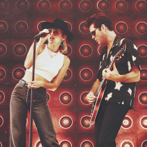 Mark Ronson and Miley Cyrus rocking out on stage. Miley, with a dynamic pose, grips the microphone, wearing a white crop top, black hat, and shades. Mark, focused on his guitar, dressed in a black and white shirt. Circular shapes and stage equipment form the backdrop.