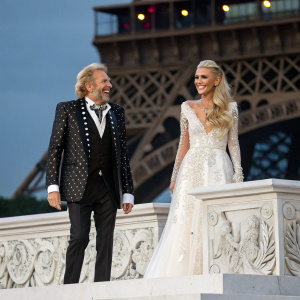 Thomas Gottschalk and Paris Hilton at the Eiffel Tower opening ceremony. They are standing on a grand marble stage with intricate designs. Both are dressed in formal attire and smiling broadly.