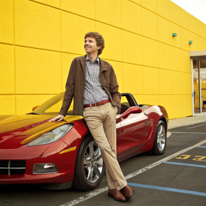 Michael Cera at a car dealership opening, posing with a shiny Chevrolet Corvette. Outdoors with a yellow painted wall in the background.