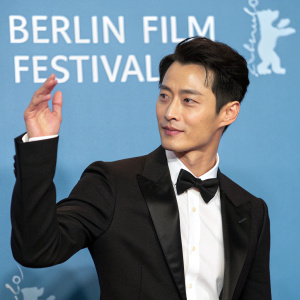 Mark Lee Ping Bin, Asian actor, attending the Berlin Film Festival award ceremony. Wearing black suit, bow tie. Dynamic pose with hand gesture. Blue background with white text. Professional photograph.
