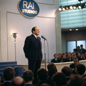 Silvio Berlusconi in a dark suit, addressing a packed auditorium at RAI Studios.