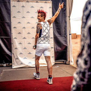 Frankie J Grande, sporting a vibrant hairstyle and an arm tattoo, stands in a candid pose. Dressed in a geometric print tank top, white shorts, and sneakers. Shot against a logo-filled black backdrop at an event.