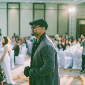 Shemar Moore, wearing a black cap and sunglasses, at a vibrant indoor gala. Diverse crowd, warm lighting, energetic ambiance