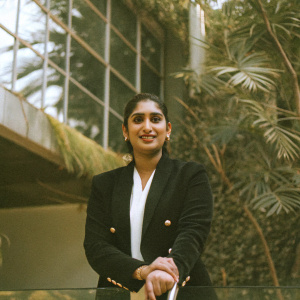 Mithali Raj, an accomplished athlete, with a radiant smile and a poised demeanor. Wearing a black blazer over a white blouse with gold buttons. The backdrop is a sleek glass building. Natural light highlights her upper body in this professional close-up portrait.