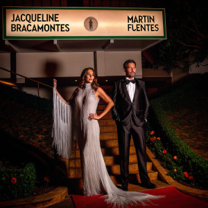 Jacqueline Bracamontes in a floor-length white gown with silver fringes. Martin Fuentes in a black tuxedo. They are posing for a photo at the gala event with white backdrop and numerous event logos visible