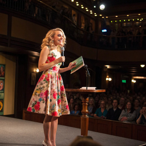 AnnaSophia Robb, wearing a floral dress, at a book launch event. Brightly lit stage with audience in the background.