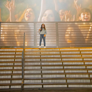 Jenna Ortega at the We Day California rally, wearing a casual outfit. Poses confidently on stage, surrounded by enthusiastic attendees.