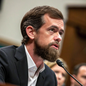 Jack Dorsey, CEO of Twitter, addressing a congressional panel. Dark suit, beard, serious expression. Background is blurred. Close-up shot of his face.