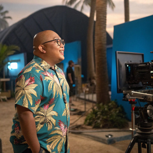 Jacob Batalon, bald and bespectacled, in a tropical shirt. On set during a movie shoot.