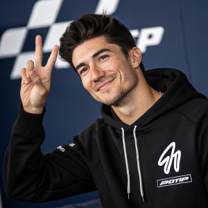 Professional photo of Maverick Viñales, MotoGP rider, casual pose, dark hair, friendly smile. Wearing black hoodie with white logo. Peace sign gesture. Dark blue backdrop with MotoGP logo