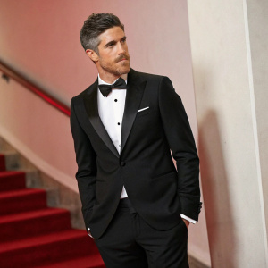 Dave Annable, actor, dressed in a sleek black tuxedo with a subtle red background, looking towards the camera at a gala event.