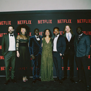 The cast of 'Sense8', a diverse group, at the Netflix awards. All wearing formal attire. Standing in front of the Netflix logo on a black backdrop. Celebratory and formal photo