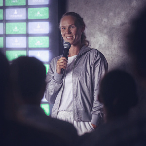 Caroline Wozniacki in a casual outfit, speaking at a tennis charity event. Neon green backdrop with sponsors logos.