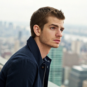 Professional headshot of Andrew Garfield. Young man with short hair, serious look, against blurred cityscape backdrop