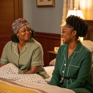 In 'LAW & ORDER: ORGANIZED CRIME', Karen Dukes as Denise and Danielle Moné Truitt as Sergeant Ayanna Bell sit on a cozy bed in a bedroom with warm-toned colors. Both actresses wear casual attire - a patterned head wrap and a green nightshirt. They are in a relaxed pose.