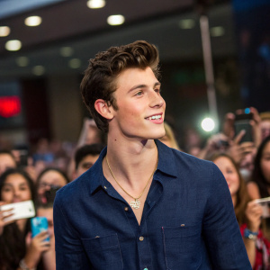 Shawn Mendes at a meet and greet event. Wearing a navy blue denim shirt and gold chain. Lively crowd in the background.