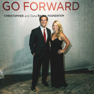 Chris Cuomo in a black suit with white shirt and red tie, and Christina Cuomo in a long black dress, posing at the 'Go Forward' charity gala sponsored by the Christopher and Dana Reeve Foundation. White backdrop with bold text and logos.
