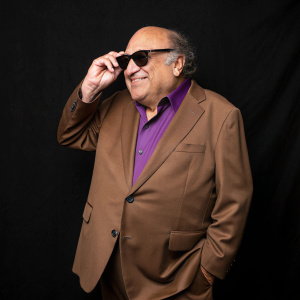 Danny DeVito, in a brown suit and purple shirt, at the BAFTA Film Awards. Adjusting sunglasses, posing against a plain black background.
