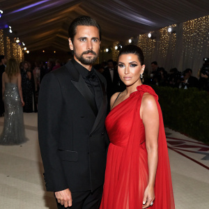 Scott Disick and Kourtney Kardashian posing at the Met Gala. He in a black suit, she in a flowing red dress. Crowded venue, dim lighting, glitzy decorations.