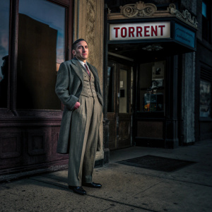 Ricardo Cortez, standing in a vintage 1920s setting, wearing a tailored suit. He holds a cigarette holder. The background is an old movie theater marquee advertising the film 'Torrent'