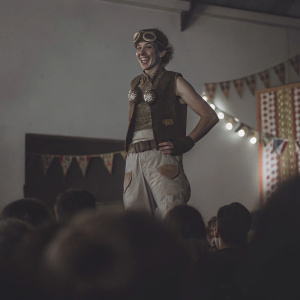 Cosplayer Hanari Solomon as Overwatch character Junkrat. Stands confidently on stage during a gaming convention. Audience and banners visible in the background.