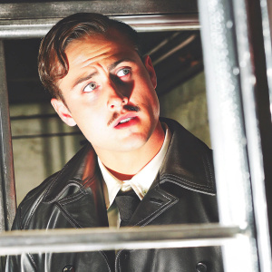 Close-up portrait of Dacre Montgomery with a mustache. Wears a black leather jacket. Dark and moody atmosphere, sharp focus on face