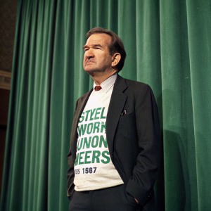 Pat Buchanan in a green curtain backdrop, wearing a dark suit with a political message t-shirt. Caucasian male, advocating for steelworkers and union members.