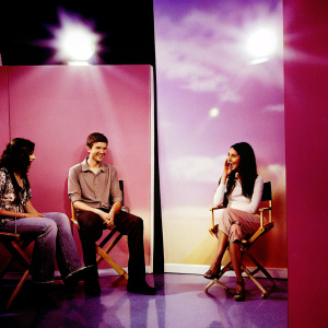 Topher Grace, Mila Kunis, and a mystery guest star in a split-screen interview setup. Colorful studio backdrop