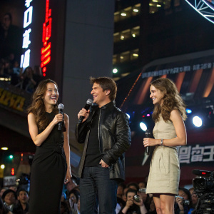 Tom Cruise, Maggie Q, and Keri Russell at a high-energy press conference for 'Mission Impossible III'. They stand on a stage with flashing lights and a cheering crowd in the background.