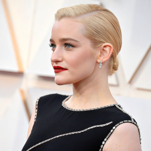 Close-up shot of Julia Garner, Caucasian actress with fair skin, blonde hair tied back, wearing a sleek black dress with subtle silver accents, red lipstick, against a soft white background.