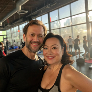 Shawn Weatherly and Jennifer Tilly at a bustling gym, natural daylight streaming through large windows, they're posing for a candid shot.
