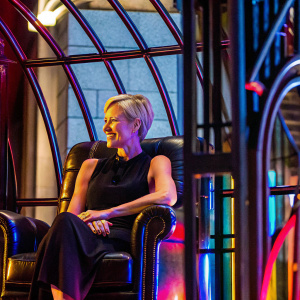 Portia de Rossi, sitting gracefully in a black leather armchair. Wearing a simple black dress and a short blonde haircut. Warm smile