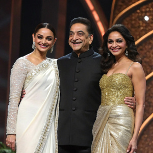 At the IIFA Awards ceremony, Kamal Haasan, Pooja Kumar, and Andrea Jeremiah pose together, all beaming with joy. Kamal Haasan, dressed in an elegant sherwani, Pooja Kumar in a white and silver sari, and Andrea Jeremiah in a gold sequined gown. The stage backdrop features a deep brown texture.