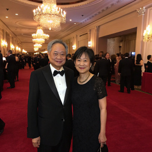 Ang Lee and Jane Lin attending a gala event. Elegant venue with chandeliers and red carpet. Ang Lee in a black tuxedo, Jane Lin in a black formal dress. Crowded room with guests.