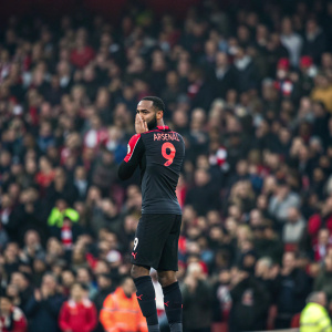 Alexandre Lacazette, wearing Arsenal's black and red jersey, number 9 clearly visible. Standing in the center of a crowded stadium, the faces of spectators a blur. His dark skin and distraught expression tell a story of profound disappointment as he covers his face.