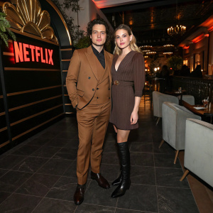 Brando Pacitto and Sofia Viscardi at a chic after party. Brando in a stylish brown suit. Sofia in a chic brown dress and high boots. The venue is adorned with Netflix branding.