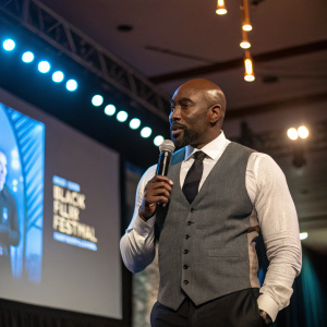 Morris Chestnut, engaged in conversation at the 2023 Black Film Festival. Formal attire, gray vest and white shirt with black tie. Bright indoor lighting, large screen behind him.
