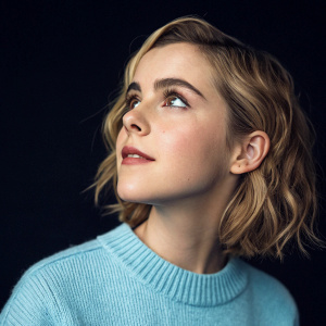Close-up of Kiernan Shipka. Soft lighting, dark background, wearing a pastel blue sweater.